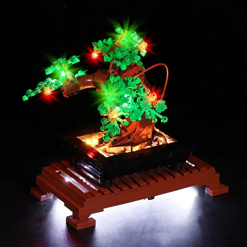 Detailansicht des beleuchteten LEGO Bonsai-Baums (Set 10281) von vorne. Der Fokus liegt auf dem Zusammenspiel der roten und grünen LED-Lichter im Laub und der warmweißen Ausleuchtung der kleinen Steine innerhalb der schwarzen Pflanzschale.