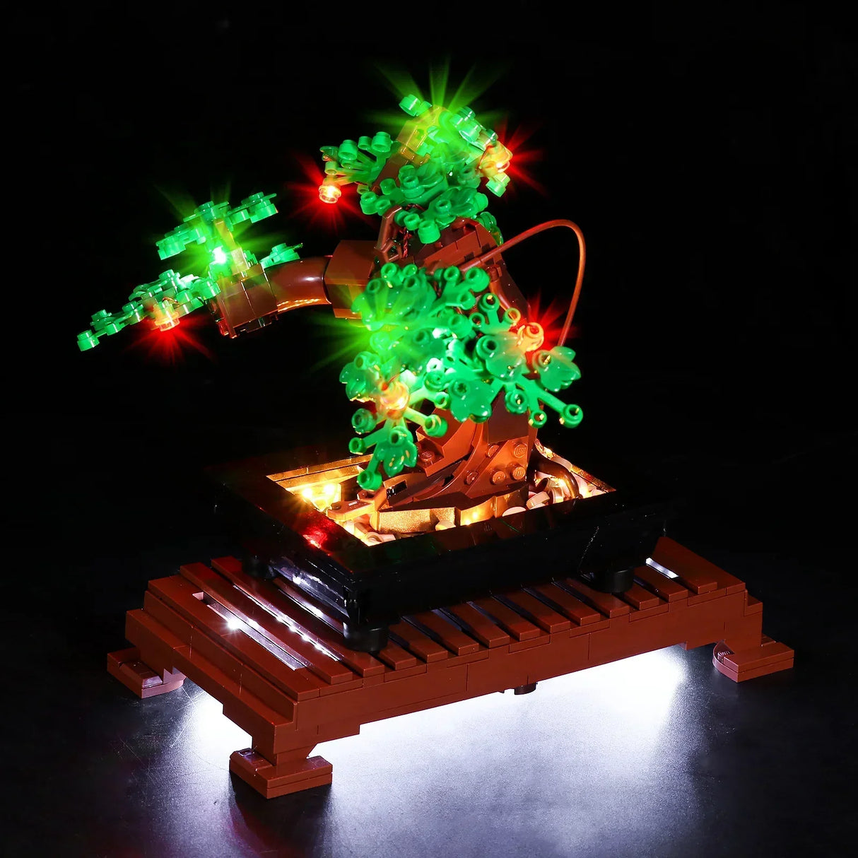 Frontale Nahaufnahme des LEGO Bonsai-Baums mit LED-Beleuchtung vor schwarzem Hintergrund. Gut sichtbar ist der geschwungene, braune Stamm, dessen Blätterkronen durch punktuelle rote und grüne LEDs intensiv leuchten. Das Licht unter dem braunen Holzgitter-Sockel erzeugt eine starke Reflexion auf der Standfläche.