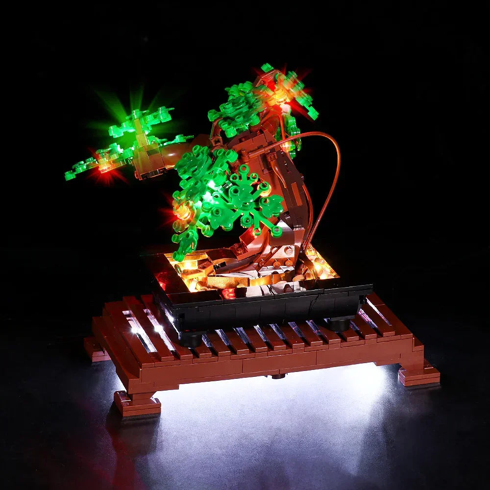 Perspektivische Schrägansicht von vorne links auf den LEGO Bonsai-Baum mit grünem Laub. Die LED-Beleuchtung setzt die Blätterkronen mit roten und grünen Lichtern in Szene. Warmes Licht strahlt aus der Schale nach oben und wirft einen hellen Schein durch das braune Podest nach unten auf den Boden.