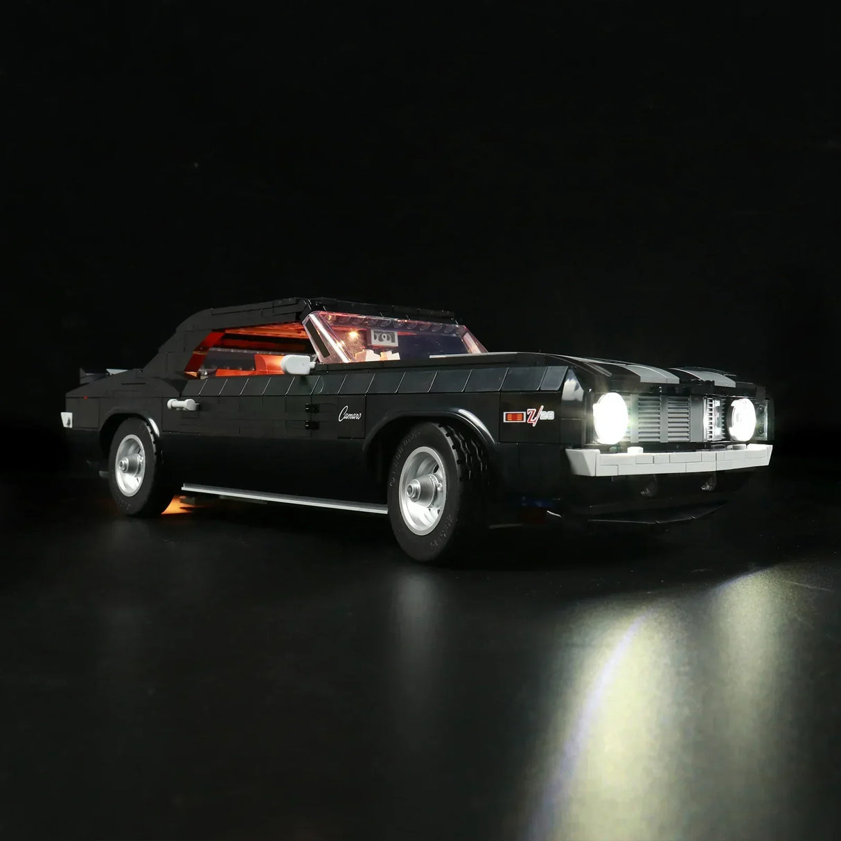 Schräge Frontansicht des schwarzen LEGO Chevrolet Camaro Z28 Modells mit LED-Licht-Upgrade. Die hellen Scheinwerfer werfen einen Lichtkegel auf den Boden, der Innenraum leuchtet rot.