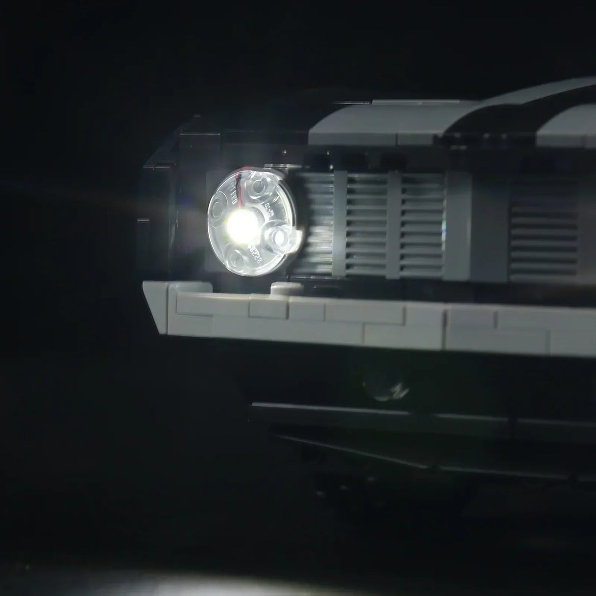 Detailaufnahme des vorderen LED-Scheinwerfers am LEGO Chevrolet Camaro Z28. Das kaltweiße LED-Licht strahlt hell durch den transparenten runden LEGO-Stein neben dem Kühlergrill.