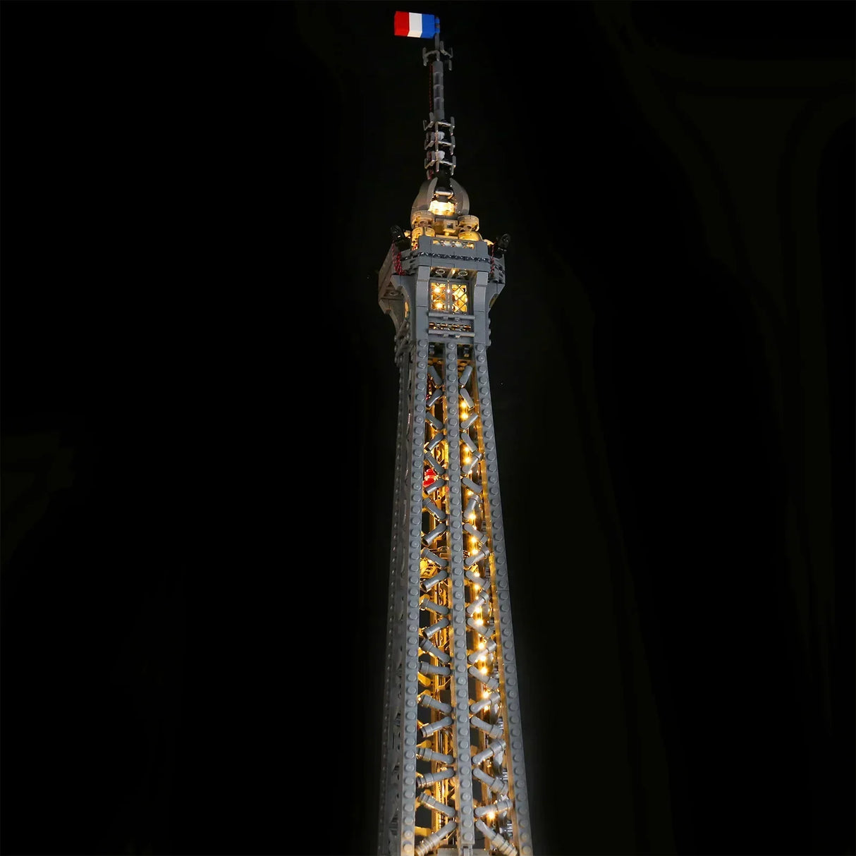 Nahaufnahme der beleuchteten Spitze des LEGO Eiffelturms 10307 mit warmer LED-Beleuchtung und französischer Flagge vor dunklem Hintergrund. 44883432702180