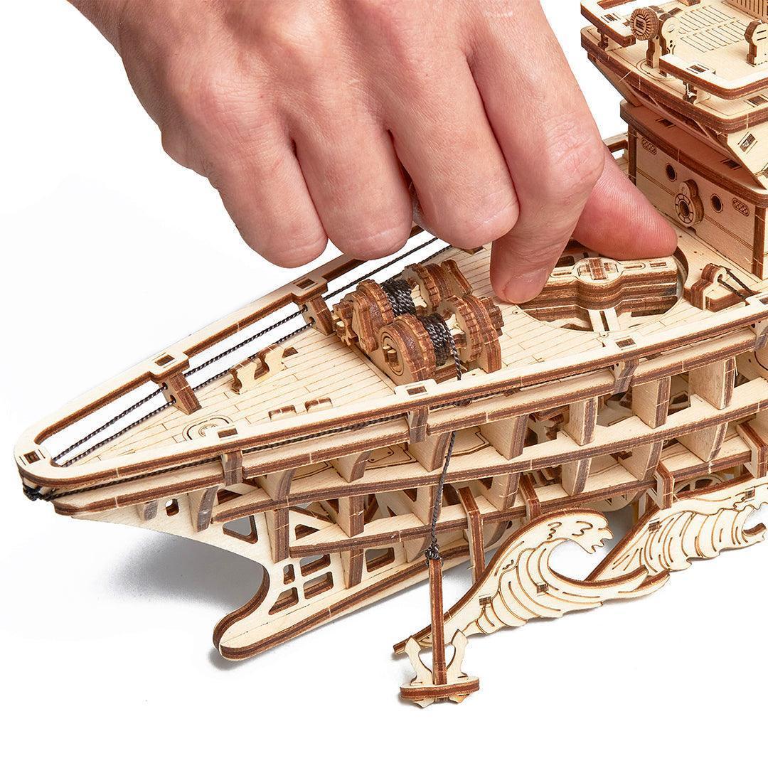 Wood Trick Forschungsschiff 3D-Bausatz mechanisch – Nahaufnahme einer Hand, die das hölzerne Zahnradgetriebe auf dem Schiffsdeck bedient.